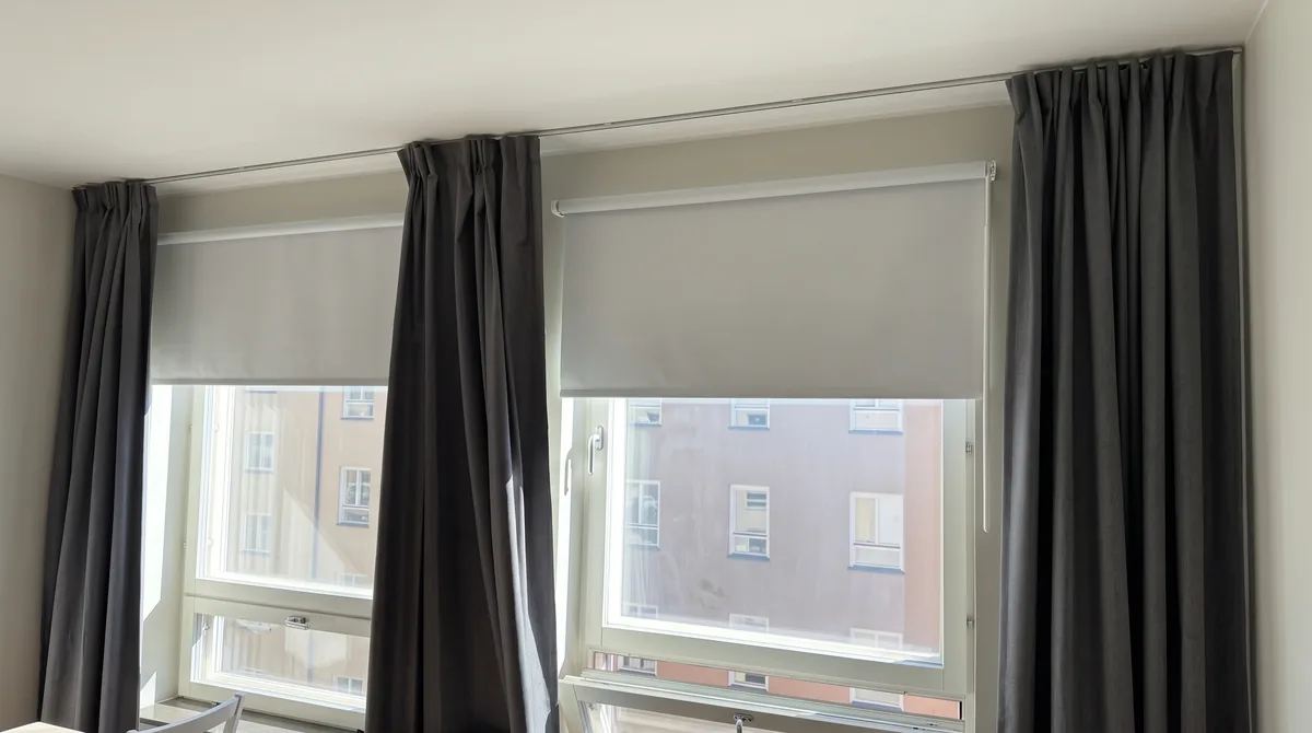 Mörkgrå gardiner på en gardinstång monterad i taket framför två fönster med ljusa rullgardiner